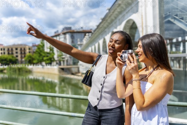 Two female tourists capturing photos and enjoying the vibrant cityscape by the river during a sunny summer vacation