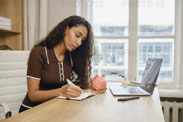 A young woman sits at a wooden desk, diligently writing in her notebook while referring to her laptop. A pink piggy bank sits nearby, indicating a focus on budgeting and savings