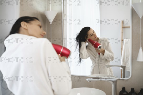 In a stylish bathroom, a woman in a white robe uses a hairdryer to dry her wet hair. Sunlight streams through a window, creating a warm and inviting atmosphere as she prepares for her day
