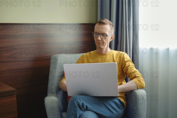 Middle-aged man in a yellow shirt sits comfortably in a grey armchair, focused on his laptop. Natural light filters through the window, creating a warm and inviting atmosphere for productivity