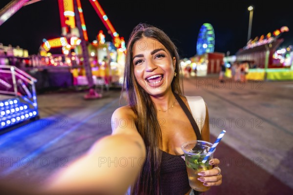 Happy young woman taking a selfie while enjoying a refreshing drink at a vibrant amusement park at night