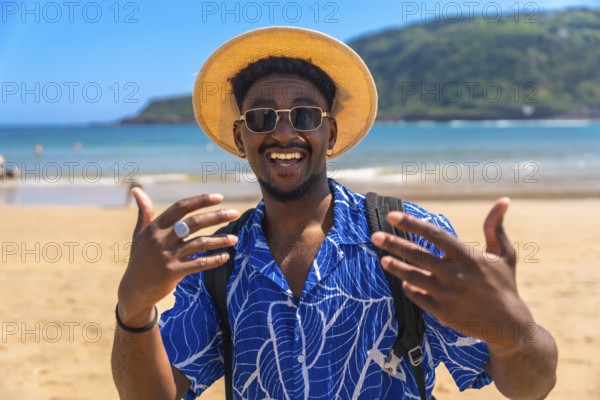Young man wearing a hat and sunglasses, joyfully gesturing with his hands while enjoying the sunny tropical beach and ocean views