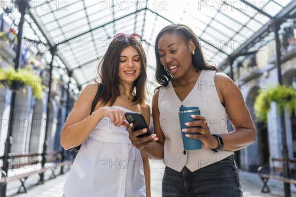 Two happy businesswomen walking through a city gallery, sipping coffee and using a smartphone, enjoying their time together outdoors