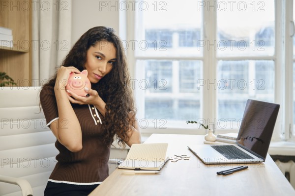 Cute woman with long curly hair is smiling and holding a pink piggy bank. She is seated at a stylish desk with a laptop and an open notebook, surrounded by large windows letting in natural light