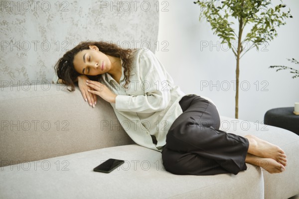 A young woman is comfortably curled up on a light-colored couch, peacefully napping. Soft sunlight filters into the cozy living room, creating a serene atmosphere