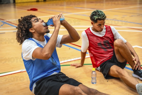 Two basketball players are sitting on the court, taking a break and drinking energy drinks after an intense training session
