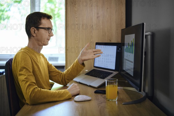A middle-aged man in a yellow shirt gestures with his hand while presenting data on a monitor in a home office