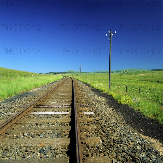 The railway track stretches through a lush green rural area, flanked by vibrant grasslands and a peaceful atmosphere. Power lines run alongside the track, enhancing the natural beauty of the scene. Cantal. Auvergne Rhone Alpes. France