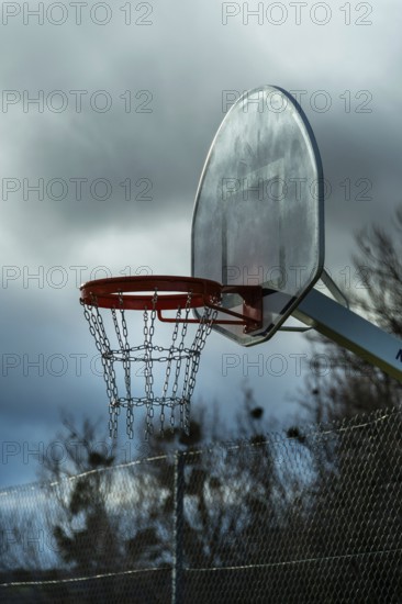 A solitary basketball hoop stands under overcast skies as dusk approaches. The chain net hangs motionless, with trees silhouetted against the darkening backdrop. France
