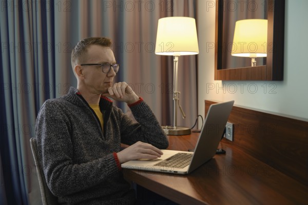 Middle-aged man sits at a wooden desk in a hotel room, focused on his laptop and is watching a sports match online. The warm lamp light creates a cozy atmosphere, while the curtains softly frame the space, suggesting evening hours