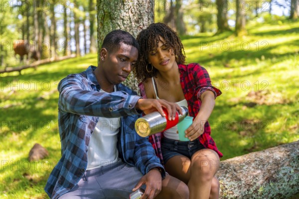 Young couple sharing a hot drink from a thermos while taking a break during their hike in a sunny forest