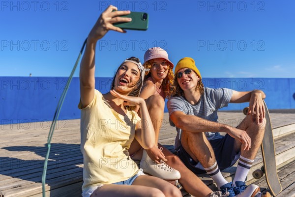 Three cheerful young friends are sitting on a wooden walkway by the sea, taking a selfie with a smartphone