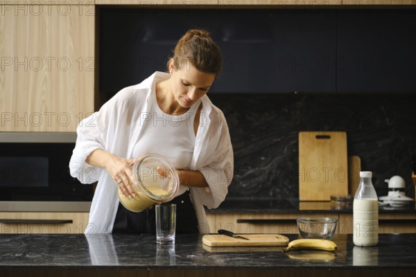 A person is preparing a healthy smoothie by pouring a blended mixture of banana and sea buckthorn into a glass. The modern kitchen is well-organized with utensils and fresh ingredients visible