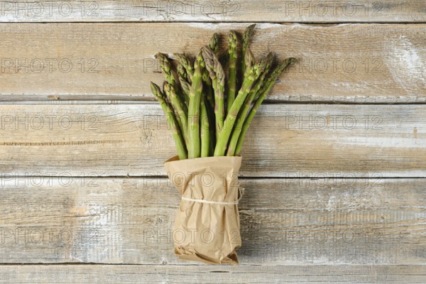 A bundle of fresh green asparagus is wrapped in brown paper and tied with twine, resting on a rustic wooden table surface highlighting its natural texture and color