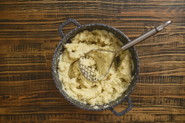 Creamy mashed potatoes are freshly prepared in a pot, sitting on a rustic wooden table. A potato masher rests inside the pot, ready for serving. This cozy cooking moment captures comfort food