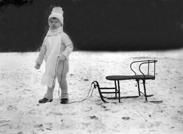 Child with sledge, 1930s, Child in winter clothes with cap and scarf standing next to a sledge in the snow, Historical photo