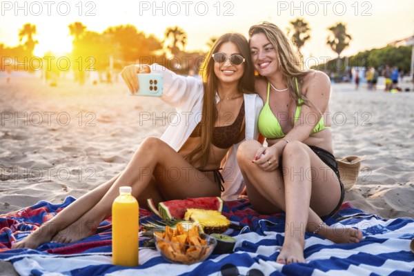 Two female friends are enjoying a beach picnic at sunset, taking a selfie with fresh fruit and juice