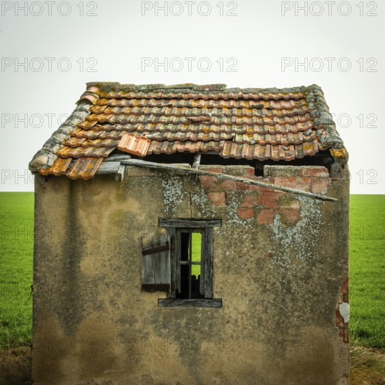 A small, abandoned structure shows signs of wear, with a crumbling roof and exposed brick. The surrounding green field highlights the building's neglect against a clear sky. Puy de Dome, Auvergne, France