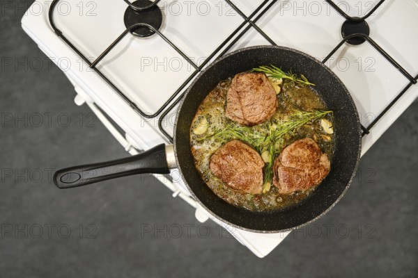 Three thick beef steaks are searing in a skillet on a gas stove. Fresh herbs and garlic surround the meat, infusing it with flavor