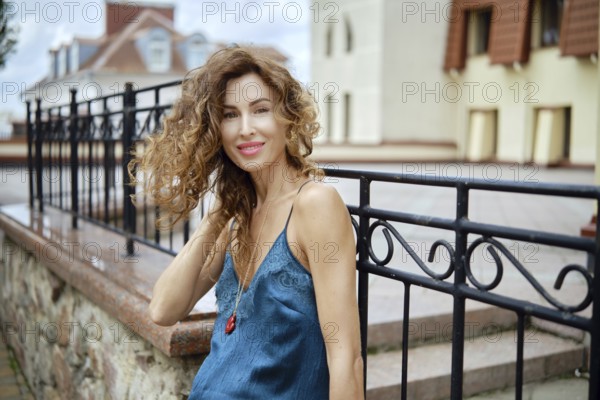 A stylish woman enjoys a relaxed moment outdoors, leaning against a stone railing while smiling at the camera. Her curly hair and blue dress complement the charming backdrop of elegant buildings