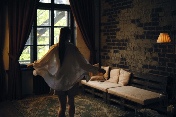 A woman in a light outfit twirls gracefully in a dimly lit room. Sunlight filters through large windows, casting shadows on a rustic sofa and creating a warm, inviting atmosphere during the evening