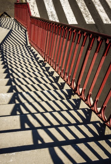 Bright sunlight creates intriguing patterns as shadows fall from red railings onto stone steps next to a striped crosswalk, adding depth and contrast to the urban landscape