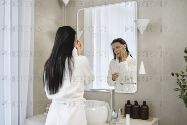 A woman in a bathrobe applies skincare products while looking at herself in a mirror