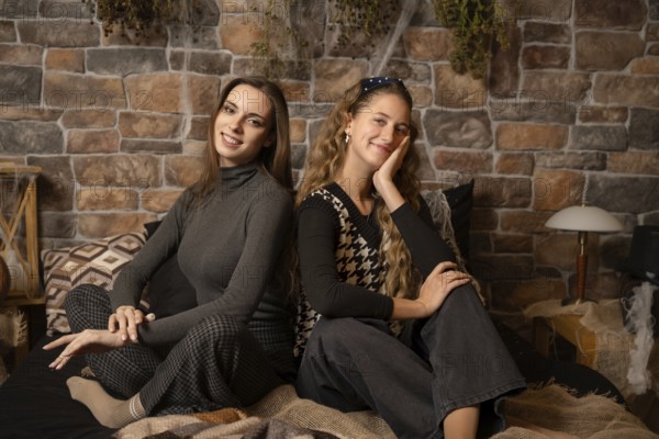 Two young women pose comfortably in a cozy indoor space featuring a rustic stone wall. The atmosphere is relaxed, with soft lighting enhancing their friendly expressions and stylish outfits