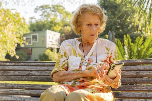 Senior woman sitting on a wooden bench in a sunny park, using a mobile phone to stay connected while enjoying the warm weather and the beauty of nature around her