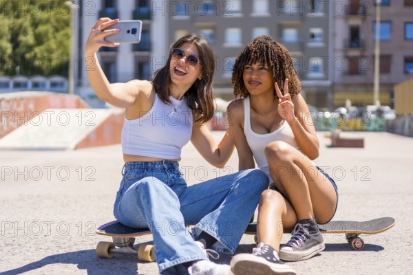 Two young women are taking a selfie with a smartphone while sitting on their skateboards in a skate park, enjoying a sunny day
