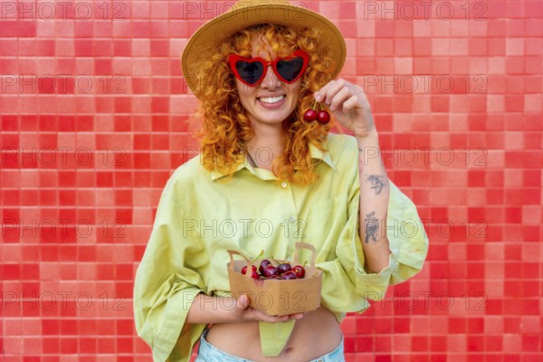 Happy young woman with curly red hair eating cherries from a small cardboard box, wearing heart shaped sunglasses and a straw hat, standing against a red tiled wall