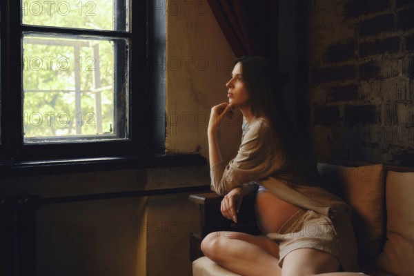 A pregnant woman sits on a couch in her home, looking thoughtfully out of a window. The afternoon light streams in, adding warmth to the cozy atmosphere indoors
