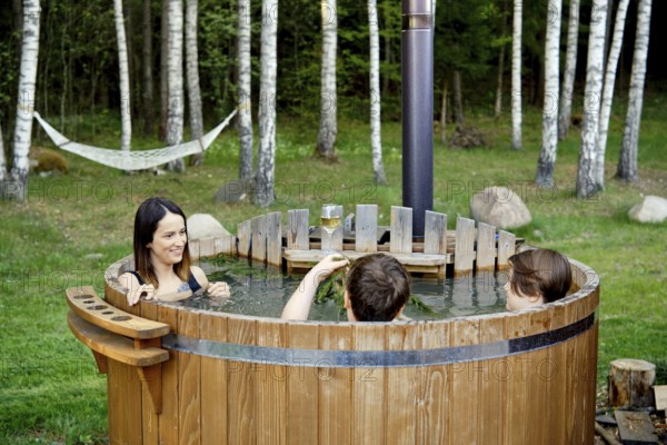 Two children and an their mom enjoy a warm soak in an outdoor wooden hot tub. Lush green grass and tall trees create a peaceful atmosphere perfect for relaxation and bonding