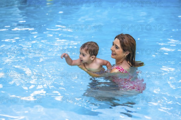 Happy mother holding her toddler son while teaching him to swim in a pool with clear blue water