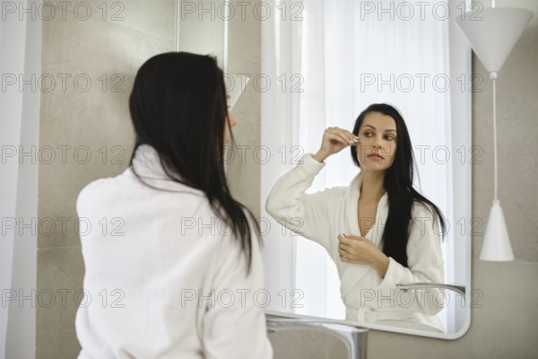 A woman dressed in a white robe stands before a mirror, carefully applying moisturizing serum in a bright bathroom. Soft morning light filters through the window, creating a serene atmosphere
