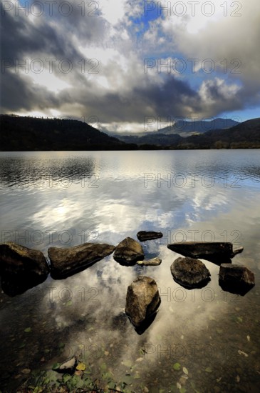 Serene waters reflect the cloudy sky at Lake Chambon, surrounded by mountains in Auvergne-Rhone-Alpes, France. The rocky shoreline adds a natural touch to this picturesque setting