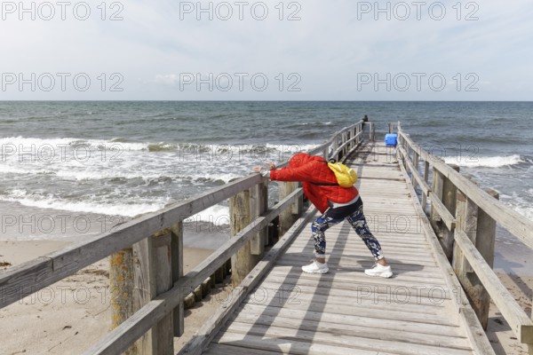 Woman in padded jacket doing stretching exercise with lunge on a wooden walkway on the beach, Baltic Sea near Kühlungsborn, Mecklenburg-Vorpommern, Germany