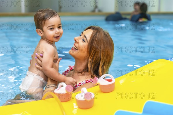 Mother holding her smiling son in a swimming pool next to floating toys, enjoying a playful moment together