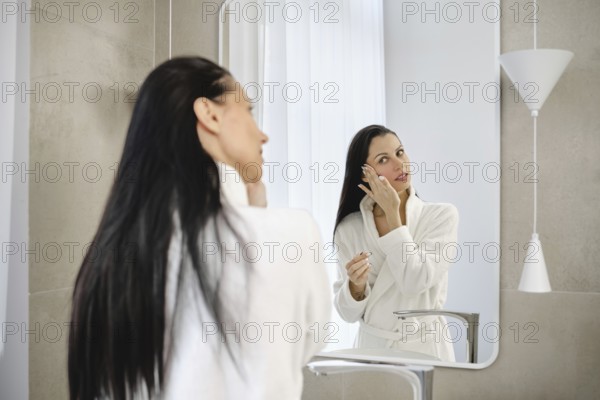 In a stylish bathroom, a woman in a white robe washing off makeup in front of the mirror. She appears focused and serene in the well-lit, contemporary environment