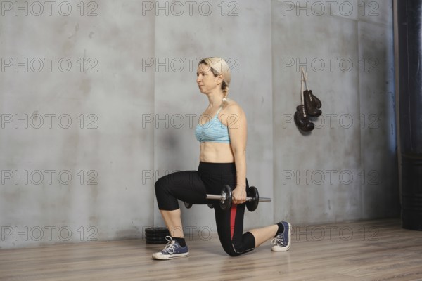 A woman with blonde hair demonstrates proper form while doing lunges with a dumbbell. She exercises in a well-lit gym with wooden flooring, focusing on strength training and fitness