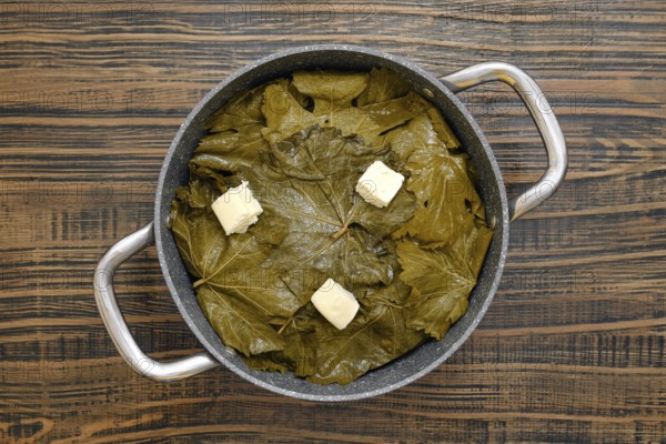 A pot filled with grape leaves prepares to cook dolma on a wooden table. Butter sits on top, ready to melt and enhance the flavors. The kitchen showcases a warm, inviting atmosphere