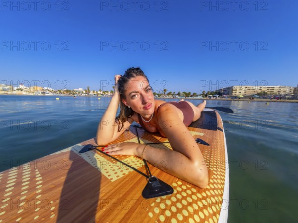 Young woman lying on a paddle board, soaking up the warm summer sun while enjoying the calm, serene sea and beautiful blue sky
