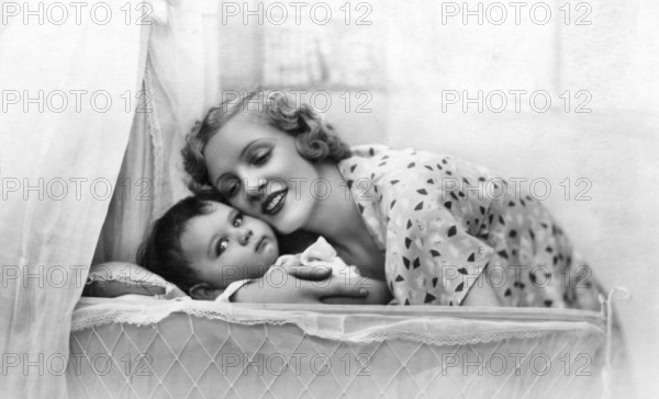 Mother embracing child, 1920s, Smiling mother bends over a baby in a cradle, surrounded by light curtains, Historical photograph