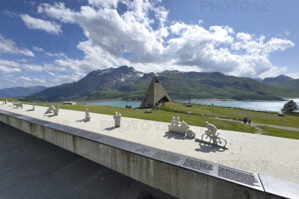 View from viewing platform to Mont-Cenis Pyramid Museum with stylised pyramid as museum in memory of Egyptian campaigns of Emperor Napoleon Bonaparte and time in front of construction of reservoir, behind it Lac du Mont Cenis, in front on parapet small stylised figures cyclists motorcyclists hikers, Mont Cenis Massif, Massif du Mont-Cenis, Graian Alps, Lanslebourg, Rhone-Alpes, France