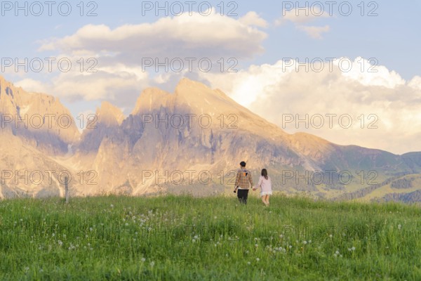 A couple walks across a flowering meadow towards the mountain range at sunset, Alpe di Siusi, Dolomites, South Tyrol, Italy