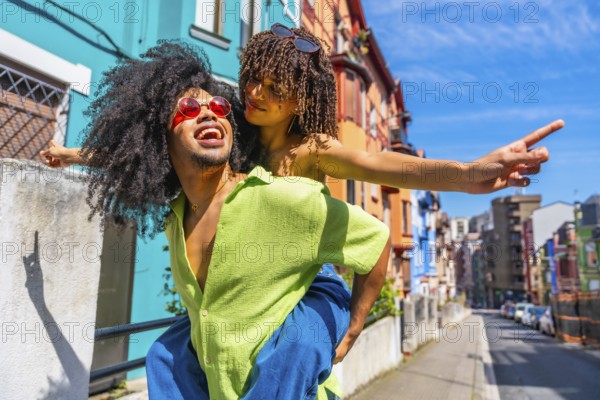 Happy tourists having fun piggybacking in a colorful city street, enjoying summer holidays and pointing at something