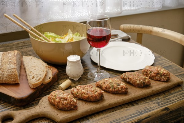 A cozy dining setup features savory meat patties served on a wooden board alongside a fresh salad in a bowl and a glass of red wine. The warm atmosphere is enhanced by the soft natural light