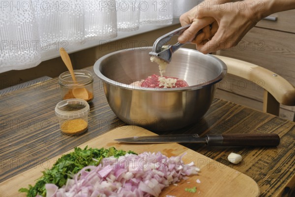Hands mincing garlic into a metal bowl filled with ground meat while chopped onions and fresh herbs lay on the wooden cutting board. The kitchen has natural light coming through