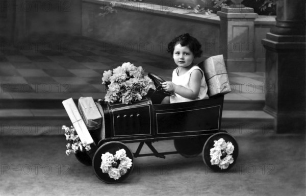 Child in toy car, 1920s, A child drives a toy car, decorated with flowers, in a nostalgic black and white photograph, Historical photograph