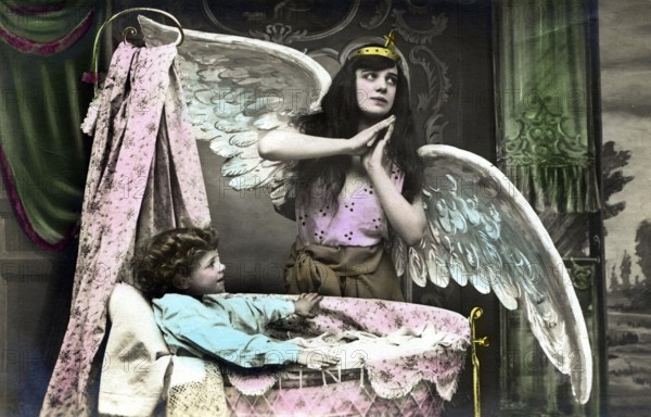 Angel with infant, 1920s, child lying in a cradle while a girl poses as an angel, historical photo